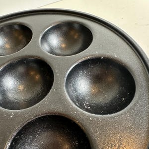 May include: Close-up of a black cast iron pan with multiple round indentations. The pan is circular with a raised edge and is designed for cooking small, round items. The surface has a slightly textured appearance.