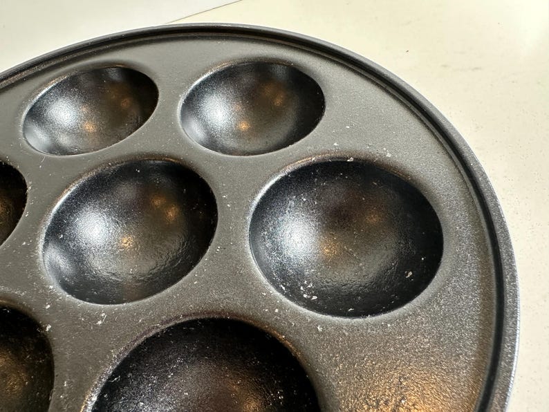 May include: Close-up of a black cast iron pan with multiple round indentations. The pan is circular with a raised edge and is designed for cooking small, round items. The surface has a slightly textured appearance.