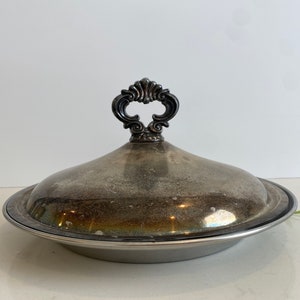 Puede incluir: Un plato de servir de plata con tapa y un asa decorativa. El plato tiene un acabado ligeramente deslustrado.