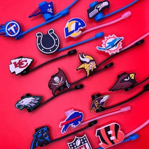 May include: A collection of NFL team logo charms attached to flexible silicone straps in various colors including blue, black, and pink. The charms feature iconic football team emblems and the NFL shield logo, designed for attachment to footwear or accessories.