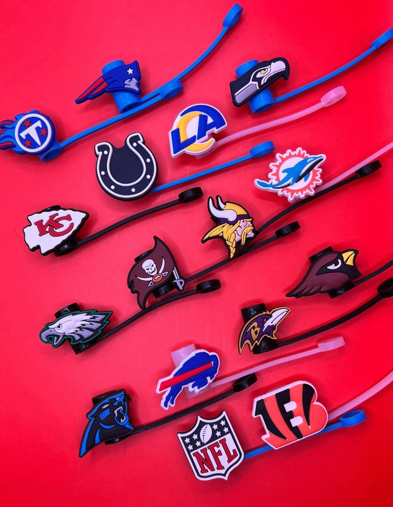 May include: A collection of NFL team logo charms attached to flexible silicone straps in various colors including blue, black, and pink. The charms feature iconic football team emblems and the NFL shield logo, designed for attachment to footwear or accessories.