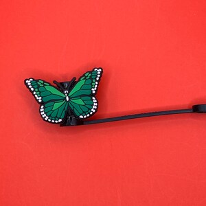 Pu&ograve; includere: Un ciondolo a forma di farfalla verde con dettagli bianchi attaccato a un cordino nero. Il ciondolo &egrave; un accessorio decorativo per una custodia per telefono o una borsa.