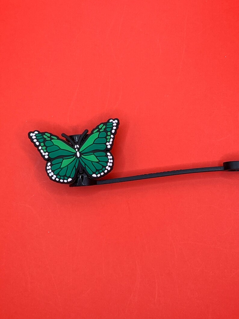 Pu&ograve; includere: Un ciondolo a forma di farfalla verde con dettagli bianchi attaccato a un cordino nero. Il ciondolo &egrave; un accessorio decorativo per una custodia per telefono o una borsa.