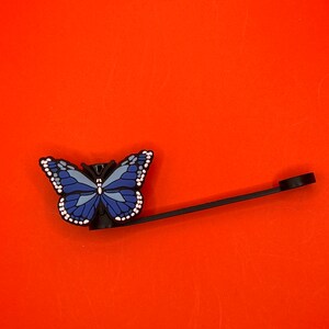 Pu&ograve; includere: Un tappo per bottiglia a forma di farfalla blu con un anello di metallo nero. La farfalla ha accenti bianchi ed &egrave; attaccata a un anello di metallo nero.