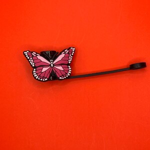 Pu&ograve; includere: Un tappo per bottiglia a forma di farfalla nero e rosa con uno spago nero attaccato.