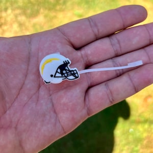 May include: A white football helmet charm featuring a yellow lightning bolt graphic on the side and a black face mask. The charm is attached to a thin, flexible white plastic stem, designed for insertion into footwear or similar accessories.