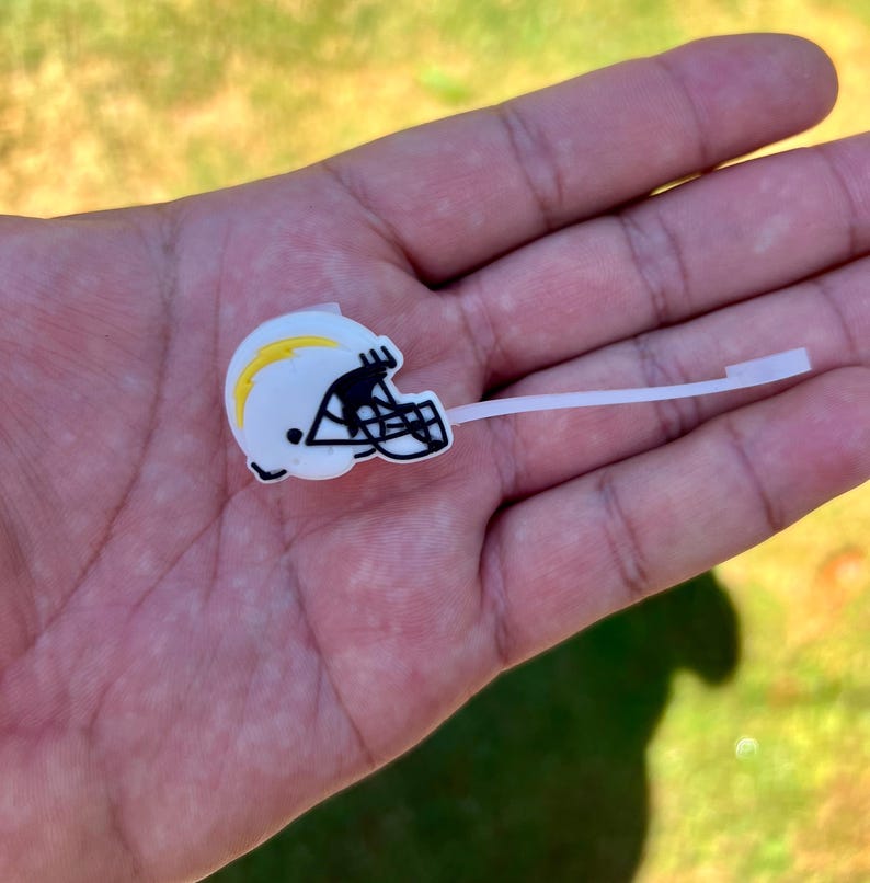 May include: A white football helmet charm featuring a yellow lightning bolt graphic on the side and a black face mask. The charm is attached to a thin, flexible white plastic stem, designed for insertion into footwear or similar accessories.