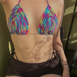 Pode incluir: Um top de biquíni de crochê colorido com uma parte inferior de amarração lateral. A parte superior é feita de um fio multicolorido com um padrão de arco-íris. A parte inferior é preta e tem um design de amarração lateral.