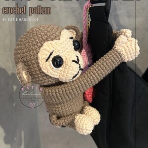 Könnte beinhalten: Ein gehäkelter Affe mit dem Text "PUNCH-KUN SNUGGIE MONKEY crochet pattern" wird gezeigt. Der Affe ist hauptsächlich braun und cremefarben, mit schwarzen Augen und einem lächelnden Gesicht. Der Affe hängt an einem schwarzen Objekt. Das Bild enthält auch den Text "34 PHOTOS".
