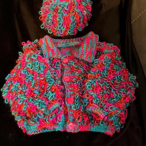 Op de afbeelding: Een handgemaakte, kleurrijke gebreide set met een jasje en een bijpassende hoed. De set is voornamelijk teal, felroze en oranje, met een textuur met lussen. Het jasje heeft een kraag en roze knopen. De hoed is rond en nauwsluitend.