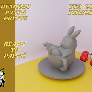 Puede incluir: Un modelo impreso en 3D gris de Pikachu sentado en una taza de té. La taza de té está sobre un platillo. Hay cuatro Poké Balls sobre una superficie amarilla. El texto "TEA-CUP PIKACHU" está en la esquina superior derecha de la imagen.