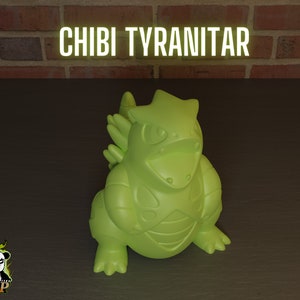 Può includere: Una statuetta di Pokémon Tyranitar verde in stile chibi. La statuetta è in piedi su una superficie marrone. Il testo "CHIBI TYRANITAR" è visibile sopra la statuetta.