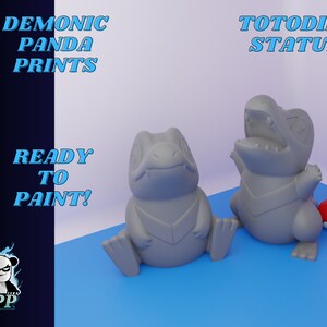 May include: Two gray Totodile statues and several red and white Poké Balls are displayed on a blue surface. The text "DEMONIC PANDA PRINTS" and "READY TO PAINT!" are visible. The image also includes the text "TOTODILE STATUE".