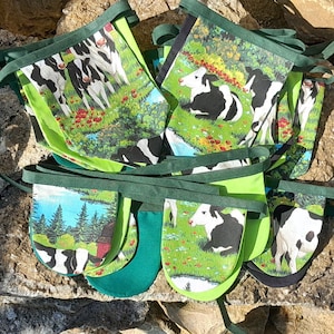 May include: A set of fabric bunting flags with a cow theme. Each flag displays a different scene of cows in a field, with green and white accents. The flags are connected by a green ribbon.