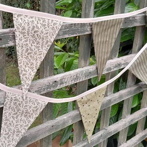 May include: A bunting banner featuring triangular flags in shades of muted green, with patterns like stripes and floral prints. The flags are strung on a pale pink ribbon and hung over a weathered wooden fence, offering a rustic, charming appearance.