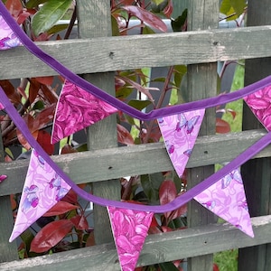 May include: A purple fabric bunting featuring a floral and butterfly design. The triangular flags are linked by a purple ribbon. The bunting is set against a weathered wooden fence and red foliage.