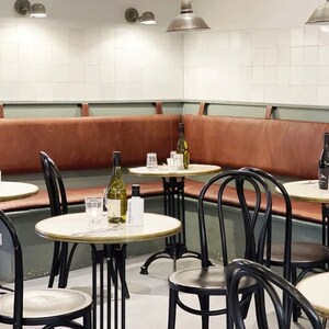 May include: A corner booth with brown leather seating and black metal chairs. There are three round tables with white tops and black metal legs. Two bottles of wine and a bottle of olive oil are on the tables.