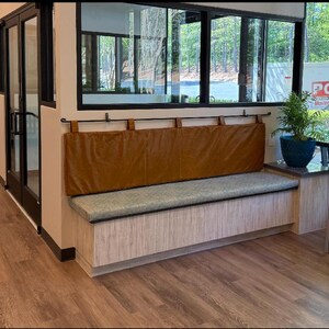 May include: A light brown leather bench with a light gray cushion and a wooden storage compartment. The bench is in a waiting area with large windows and a potted plant.