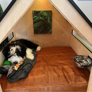 May include: A black and white dog is lying on a brown leather bed in a wooden dog house. The dog house has a small window and a green painting on the wall. There is a stainless steel dog bowl attached to the wall.
