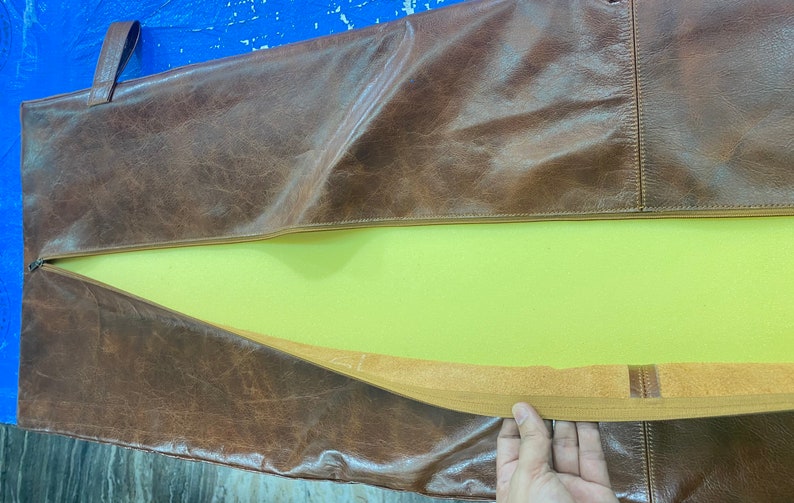 May include: A brown leather bag with a zipper opening and a yellow foam lining. The bag has a brown strap and a brown elastic band.