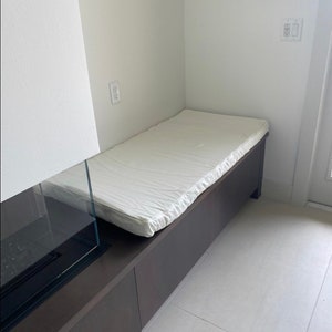 May include: A white cushioned bench with a dark brown wood base. The bench is built into a wall and has a glass fireplace on the left side.