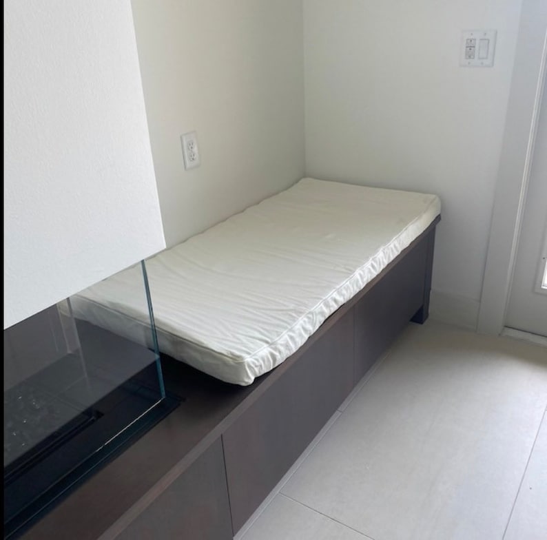 May include: A white cushioned bench with a dark brown wood base. The bench is built into a wall and has a glass fireplace on the left side.