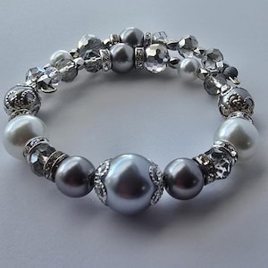 May include: A beaded bracelet featuring a mix of gray, silver, and white beads. The bracelet includes large gray pearl-like beads, smaller silver beads, and clear faceted beads. Decorative silver accents with rhinestones add sparkle.
