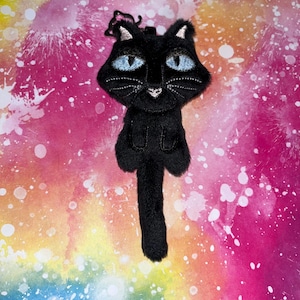 May include: A black cat plush toy with blue eyes and white accents. The cat has a long tail and is set against a colorful, abstract background. The cat's eyes are embroidered with blue thread, and it has a small, white heart-shaped nose.
