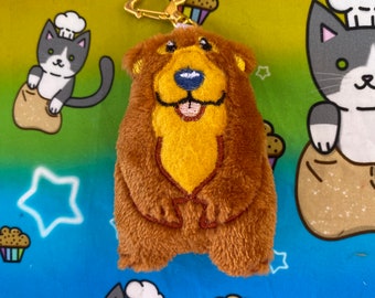 Puede incluir: Un llavero de peluche marrón en forma de oso con una cara bordada amarilla y un anillo de llave dorado. El oso lleva un sombrero de peluche marrón con un gorro de chef blanco en la parte superior.