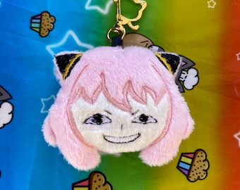 May include: A pink plush keychain featuring an embroidered Anya Forger face with a mischievous grin. The keychain has black and gold accents on the ears and a gold-colored clasp. The background is a rainbow-colored fabric with star and cupcake patterns.