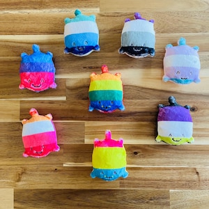 May include: A collection of nine colorful plush toys, each with a unique striped design and a smiling face. The toys feature a variety of colors, including blue, pink, purple, and rainbow stripes. They are arranged on a wooden surface.