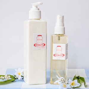 Puede incluir: Dos botellas de productos para el cuidado del cuerpo con temática de yogur. La botella más grande es una loción corporal blanca con un dispensador de bomba. La botella más pequeña es una bruma corporal transparente con una boquilla de pulverización. Ambas botellas tienen una etiqueta roja y blanca con un contenedor de yogur de dibujos animados y el texto "yogurt".