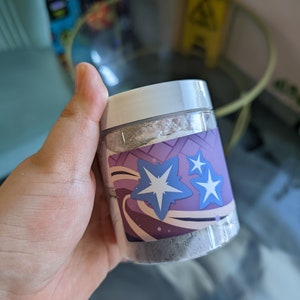 May include: A clear plastic jar with a white lid filled with a gray powder. The jar has a purple, white, and blue label with a design of three stars.