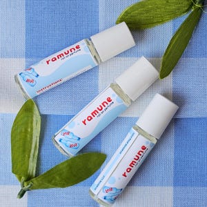 Puede incluir: Tres frascos de perfume roll-on con tapas blancas y etiquetas rojas que dicen "ramune roll on perfume". Las etiquetas tienen un diseño azul claro con ilustraciones de botellas. Los frascos están sobre una superficie a cuadros azul y blanco con hojas verdes.