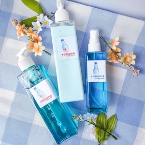 Puede incluir: Tres botellas de productos para el cuidado del cuerpo sobre una superficie a cuadros azules y blancos. Las botellas son transparentes y contienen líquido azul. Las botellas están etiquetadas como "ramune body lotion", "ramune body mist" y "ramune body wash".
