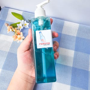 Pode incluir: Uma garrafa transparente de gel de banho azul com um dispensador de bomba branco. A garrafa tem um r&oacute;tulo com o texto "ramune body wash" em rosa e branco.