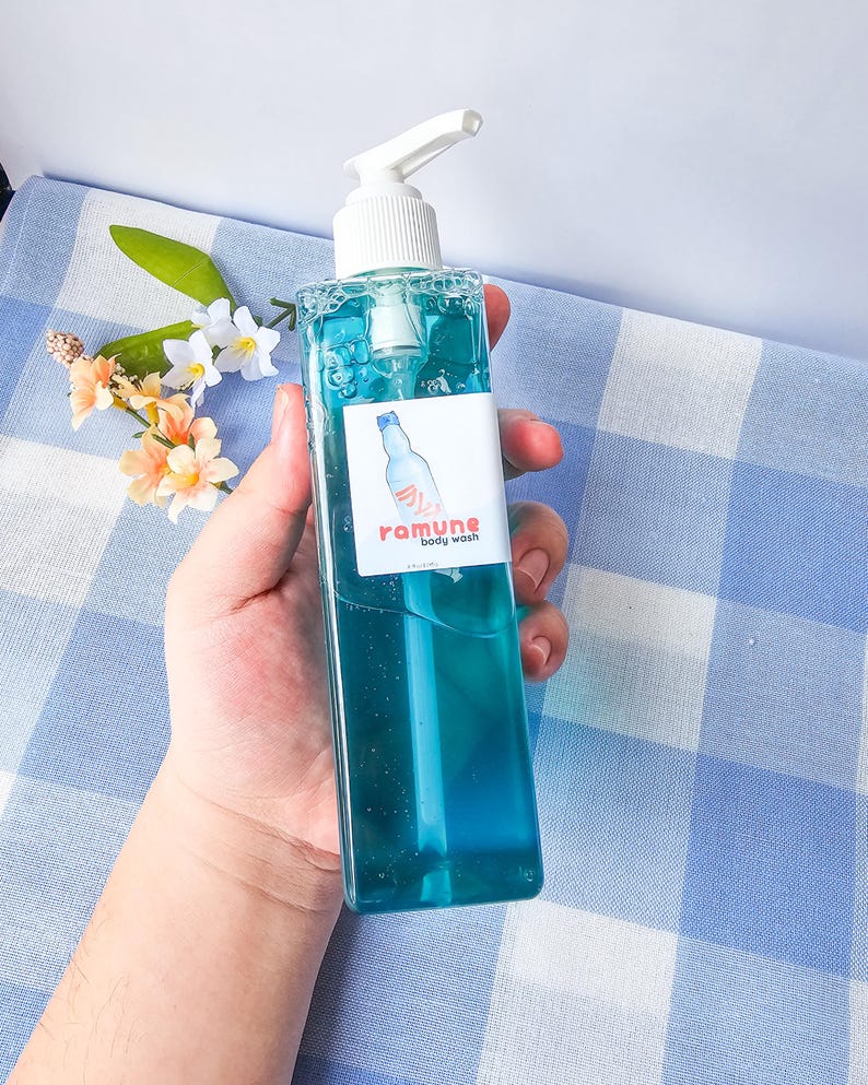 Pode incluir: Uma garrafa transparente de gel de banho azul com um dispensador de bomba branco. A garrafa tem um r&oacute;tulo com o texto "ramune body wash" em rosa e branco.