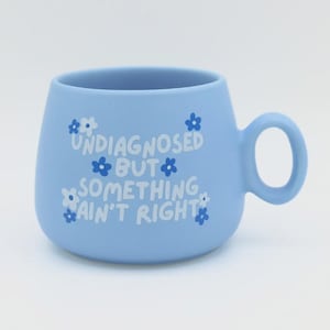 May include: A light blue ceramic mug with a rounded shape and a circular handle. The front features white and dark blue flower graphics surrounding the white text that reads: UNDIAGNOSED BUT SOMETHING AIN'T RIGHT. The mug has a smooth finish.