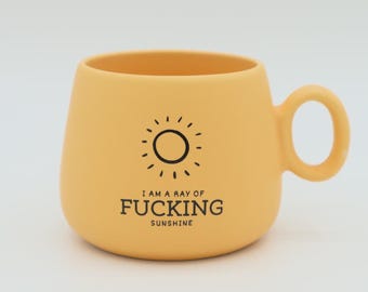 May include: A yellow ceramic mug with a rounded body and a handle. The mug features a black sun graphic and the text "I AM A RAY OF FUCKING SUNSHINE". The mug is designed for hot beverages.