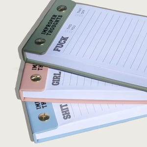 May include: Three notepads in green, pink, and blue, each with a different word printed on the cover: "FUCK", "GIRL", and "SHIT". The top notepad reads "IMPROPER THOUGHTS". Each notepad has lined pages and a metal screw closure.
