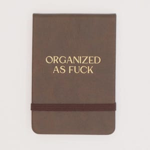 May include: A brown notebook with the text "ORGANIZED AS FUCK" in gold lettering. The notebook has a brown elastic band closure. The cover is a solid brown color.
