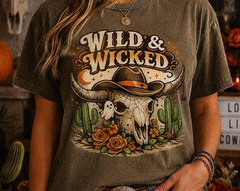May include: A charcoal gray t-shirt featuring a graphic print of a bull skull wearing a cowboy hat, surrounded by cacti, orange roses, a small ghost, and a crescent moon. The text WILD & WICKED is printed in a decorative font above the skull.