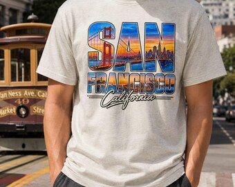 May include: A light gray heathered t-shirt featuring a graphic print of the San Francisco skyline and the Golden Gate Bridge at sunset. The text SAN FRANCISCO California is printed in bold, dark blue letters over the colorful city landscape image.