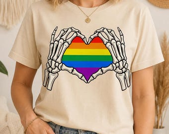 May include: A cream-colored short-sleeve t-shirt featuring a graphic print of two skeletal hands forming a heart shape. The heart is filled with horizontal stripes in the colors of the rainbow, including red, orange, yellow, green, blue, and purple.