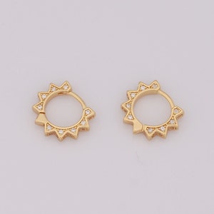May include: Two gold-colored hoop earrings. Each earring features a sunburst design with small, clear gemstones. The earrings are displayed against a plain, light background, highlighting their intricate details and design.