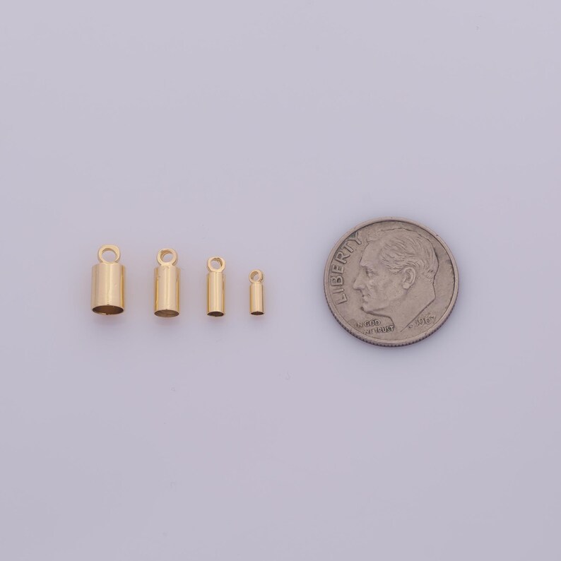 May include: Four gold-colored cylindrical jewelry findings with a loop at the top, next to a U.S. dime for size comparison. The findings vary in size, with the largest measuring approximately 0.3 inches in height.
