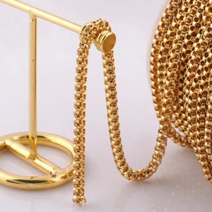 May include: A gold chain with a box link pattern. The chain is displayed on a gold stand and is coiled on a spool.