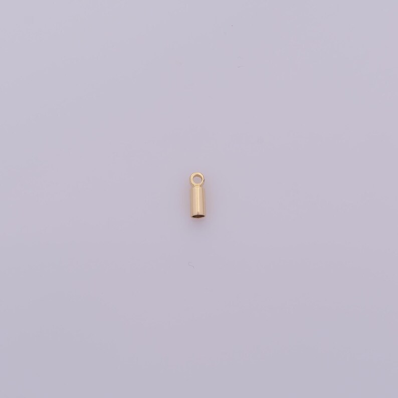 May include: A small, gold-colored jewelry finding, a crimp tube, is shown against a light purple background. The tube is cylindrical with a small loop at the top. This jewelry component is used for stringing beads and securing clasps.