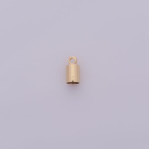 May include: A small, gold-colored cylindrical jewelry finding with a loop at the top. The finding is a cap for jewelry making, with a smooth, polished surface. The background is a light purple color.