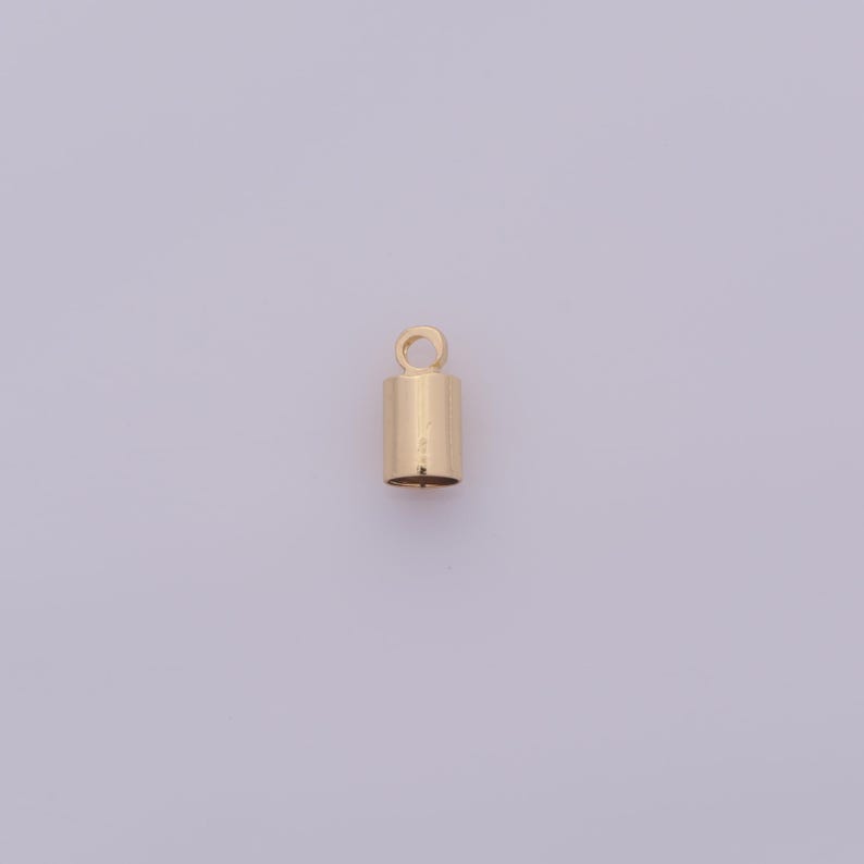 May include: A small, gold-colored cylindrical jewelry finding with a loop at the top. The finding is a cap for jewelry making, with a smooth, polished surface. The background is a light purple color.